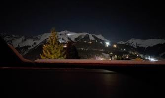 An impressive winter landscape with snow-covered mountains and lights in the distance. The sky is dark, highlighting the tranquil atmosphere.