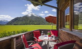 A cozy balcony with red upholstered chairs and a table. In the background, green meadows and majestic mountains can be seen under a blue sky.
