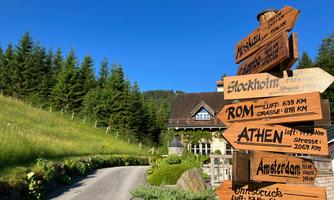 A signpost with several destinations, including Stockholm, Rome, and Athens, stands by the roadside. In the background, a beautiful house and a green, wooded landscape can be seen.