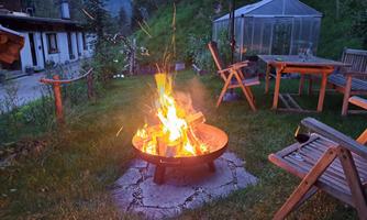 A cozy campfire in a garden with wooden chairs and a table. Surrounded by trees and a greenhouse in the background.