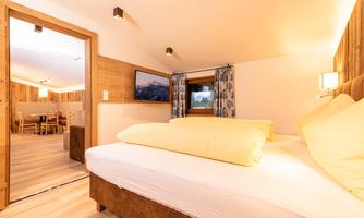 A cozy bedroom with two beds and yellow pillows. In the background, there is a dining area and a window with a view of the mountains.
