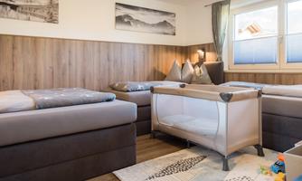 A cozy sleeping area with two single beds and a children's bed. The walls are in warm wood tones and there are large windows that let in plenty of light.