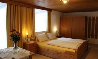 A cozy bedroom with a double bed and wooden wardrobes. The windows are decorated with light curtains and fresh flowers are on the table.