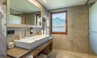 A modern bathroom with a large mirror and a sink made of white material. Through the window, you can see a beautiful mountain landscape.