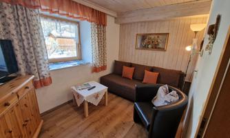 A cozy living space with a sofa, an armchair, and a wooden table. The walls are light and clad with wooden panels, and there are curtains and a television.