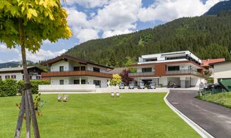 A modern residential complex with two buildings and spacious green areas. In the background, there are wooded mountains and a clear sky.