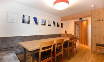 A cozy dining area with a large wooden table and several chairs. Various artworks hang on the wall, and the room is brightly lit.