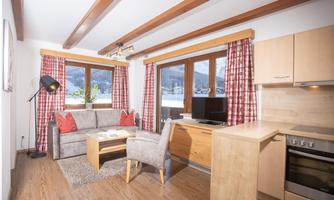 A cozy living room with a sofa, armchair, and wooden shelves. Large windows offer a view of the mountains.
