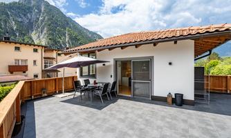 A modern terrace with a dining table and chairs, surrounded by a beautiful view of the mountains. The sky is partly cloudy and the atmosphere feels welcoming.