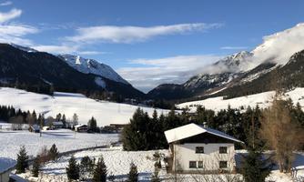 A winter landscape with snow-covered mountains and a clear blue sky. In the foreground, there is a cozy house surrounded by snow-covered trees.