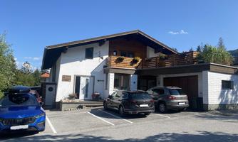 A modern house with a balcony and several cars in the parking lot. The sky is clear and there are some trees in the vicinity.