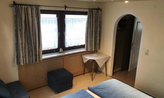 A cozy room with two beds, a small table, and a sofa. Large windows ensure plenty of daylight.