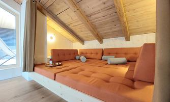 A cozy seating area with orange cushions under a wooden ceiling roof. Large windows provide plenty of light and create an inviting space.