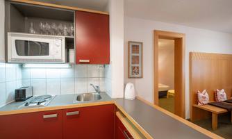 A modern kitchenette with red cabinets, microwave, and cooktops. In the background, a cozy living area with a sleeping area can be seen.