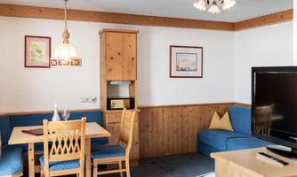 A cozy living space with wooden paneling, a dining table, and blue chairs. In the background, there is a seating area and a TV.