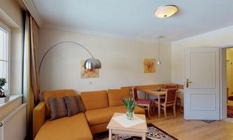 A bright living room with a comfortable orange couch and a dining table. There is a standing lamp and decorative pictures on the wall.