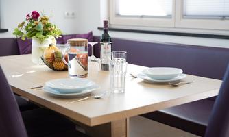 A beautifully set table with two plates, a glass, and a bottle of wine. In the background, there is a vase with flowers and fruit.