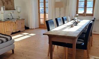A cozy dining area with a long wooden table and black chairs. Large windows let in plenty of light and offer a view outside.