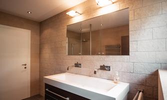 A modern bathroom with a large mirror above a minimalist sink. The walls are clad in bright stones and there is stylish lighting.