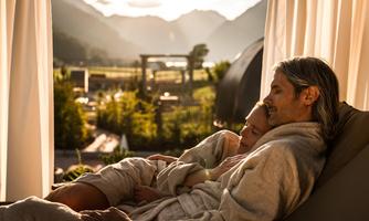 A couple is relaxing in a modern room with large windows and a view of the mountains. The atmosphere is calm and cozy, perfect for resting.