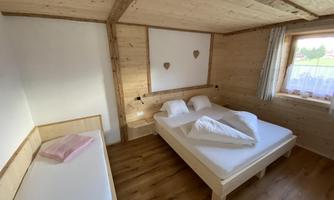 A cozy room with wooden paneling and a double bed. The room has a window with a view outside and an additional lounge chair.