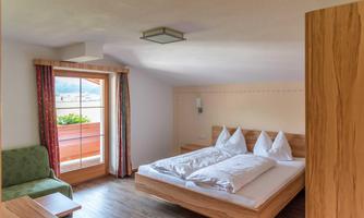 A bright, cozy bedroom with a double bed and two pillows. There is a window overlooking a balcony and a green seating area.