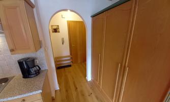A bright kitchen with wooden cabinets and a passage to a hallway. The floor is made of wood, and a coffee machine is on the countertop.