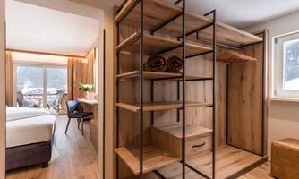 A modernly designed wardrobe with wooden shelves and elements. In the background, a cozy room with a view outside can be seen.