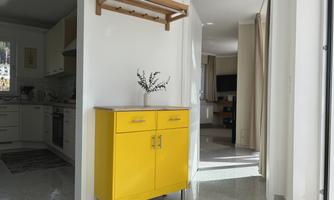 A modern room with a yellow dresser and a wooden shelf. In the background, an open kitchen and a bright space are visible.