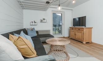 A modern living room with a comfortable sofa and colorful cushions. There is a wooden table, a TV, and large windows that let in plenty of light.