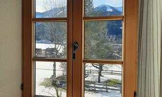 A window with a wooden frame that offers a view of a snowy landscape and mountains. On the windowsill, there are a few plants and decorations.
