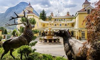 A picturesque hotel with a fountain in the foreground. Stylish animal statues are in the garden, and mountains are visible in the background.
