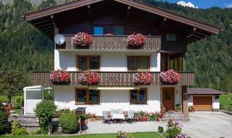 A charming three-story house in the Alps with balconies full of blooming plants. The well-maintained garden and the surrounding mountains create an idyllic landscape.