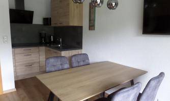 A modern kitchen unit with wooden cabinets and a dining table. The room is bright and inviting, featuring elegant chairs and modern lamps.