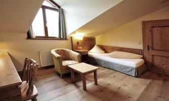 A cozy room in the attic with a window and wooden details. There is a small table, an armchair, and a bed.
