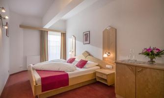 A cozy bedroom with a double bed and wooden furniture. The room is bright and friendly with windows that let in natural light.