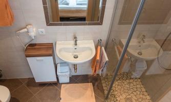A modern bathroom with a shower, a sink, and a wooden shelf. The walls are light tiled and there is a towel on a holder.
