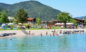A beautiful swimming lake with people swimming in the water and relaxing on the shore. In the background, green mountains and sunny lounging areas can be seen.