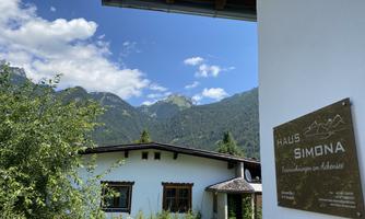 A cozy house with a sign reading "Haus Simona" in front of a picturesque mountain landscape. The sky is clear and the surroundings are green and inviting.