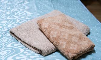 Two towels are lying on a bed with a blue bed sheet. One of the towels is light beige and the other has a subtle pattern.