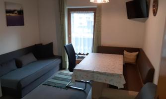 A cozy living area with a gray sofa and a table. Windows with curtains and a television on the wall complete the room.