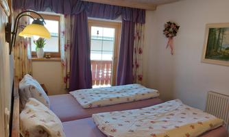 A cozy bedroom with two single beds and blooming bed covers. The room has large windows with curtains and a warm, inviting atmosphere.