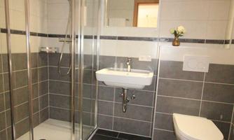 A modern bathroom with a glass shower and a sink. The floor is equipped with dark tiles, and there is a white bath mat.