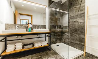 A modern bathroom with a spacious shower and a double sink. The walls are done in a gray stone look and there are fresh towels on a shelf.