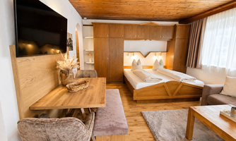 A cozy room with wooden decor, a double bed, and a television on the wall. Additionally, there is a table with chairs and plenty of daylight through the windows.