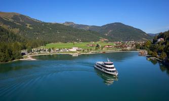 A picturesque lake with a boat, surrounded by green forests and mountains. The sky is clear and blue, perfect for a relaxed day outdoors.