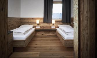 A simple room with two single beds and wooden paneling. Windows with a view of the mountains and dark curtains create a cozy atmosphere.