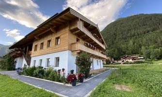 A rustic house with wooden cladding and balconies in a green landscape. In the background, mountains and a clear sky can be seen.
