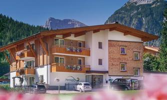 A cozy house in alpine style, surrounded by green forests and mountains. The facade is made of wood and stone with balconies.