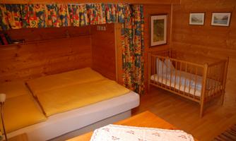 A cozy bedroom with wood paneling and a large bed. Next to the bed is a crib, and there are colorful curtains as well as some pictures on the wall.